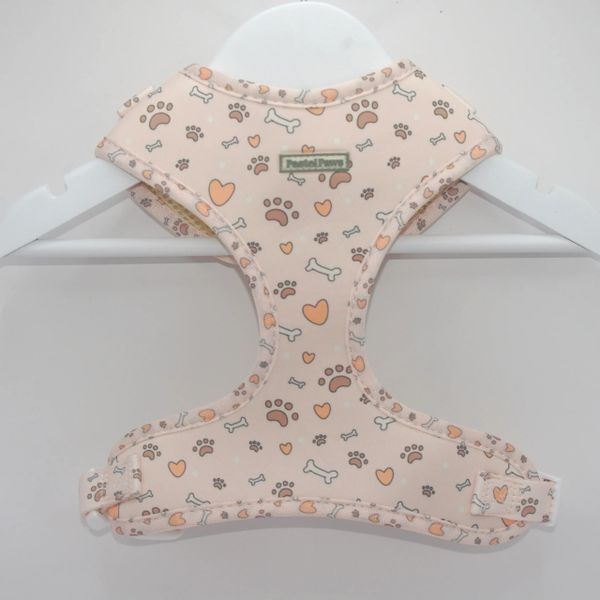 Beige dog harness with paw, bone, and heart patterns hanging on a white hanger.
