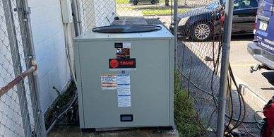 Outdoor HVAC unit installed beside a building under a chain-link fence.