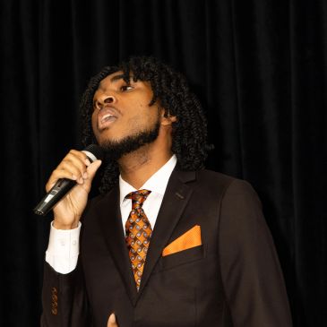 Man in suit singing into a microphone against a black background.
