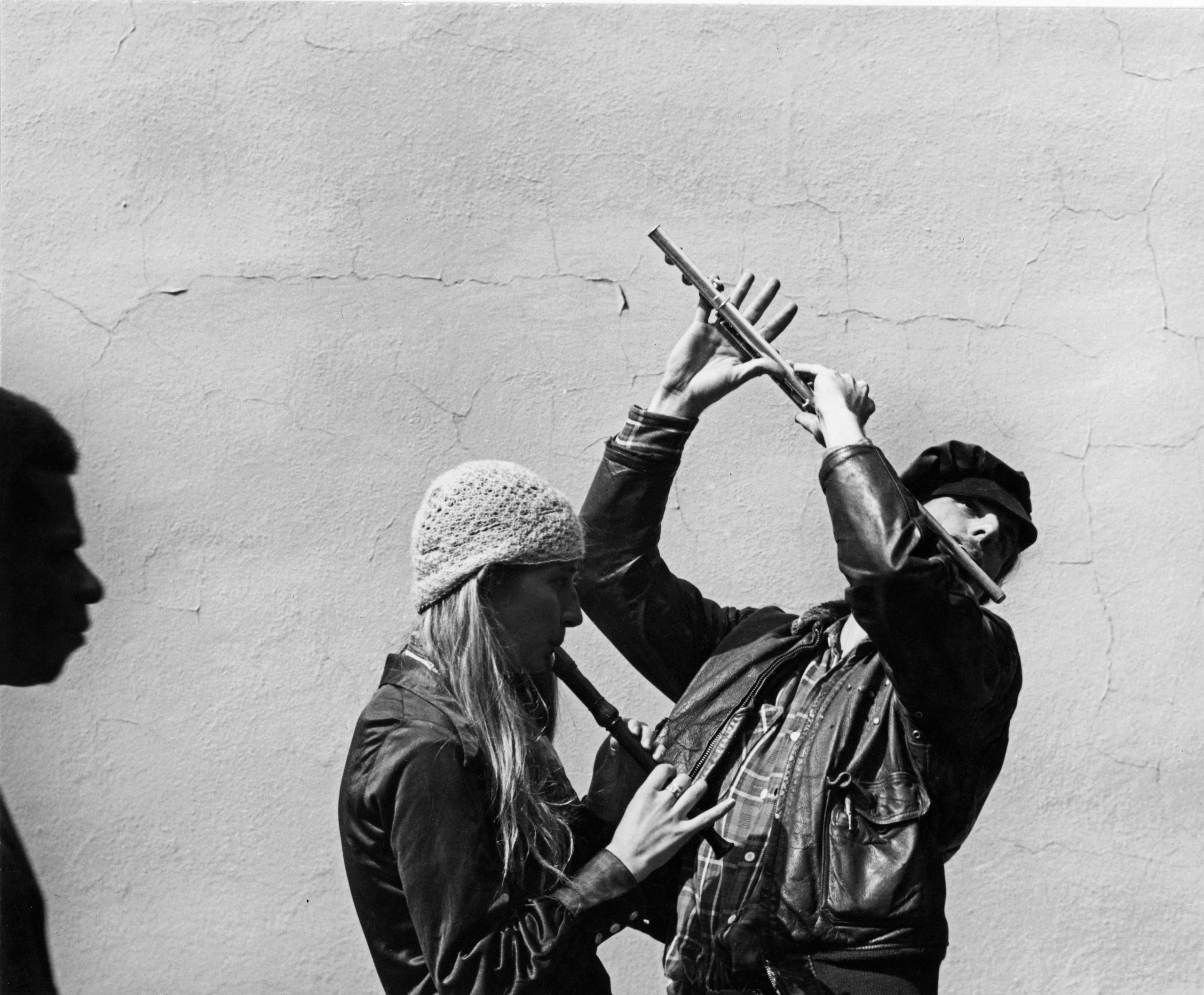 Two people playing flutes against a cracked wall.