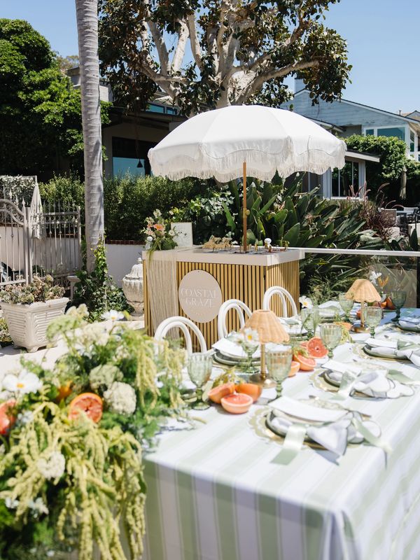 Elegant outdoor dining setup with greenery, fresh fruit, and a coastal-themed bar under a white umbrella.