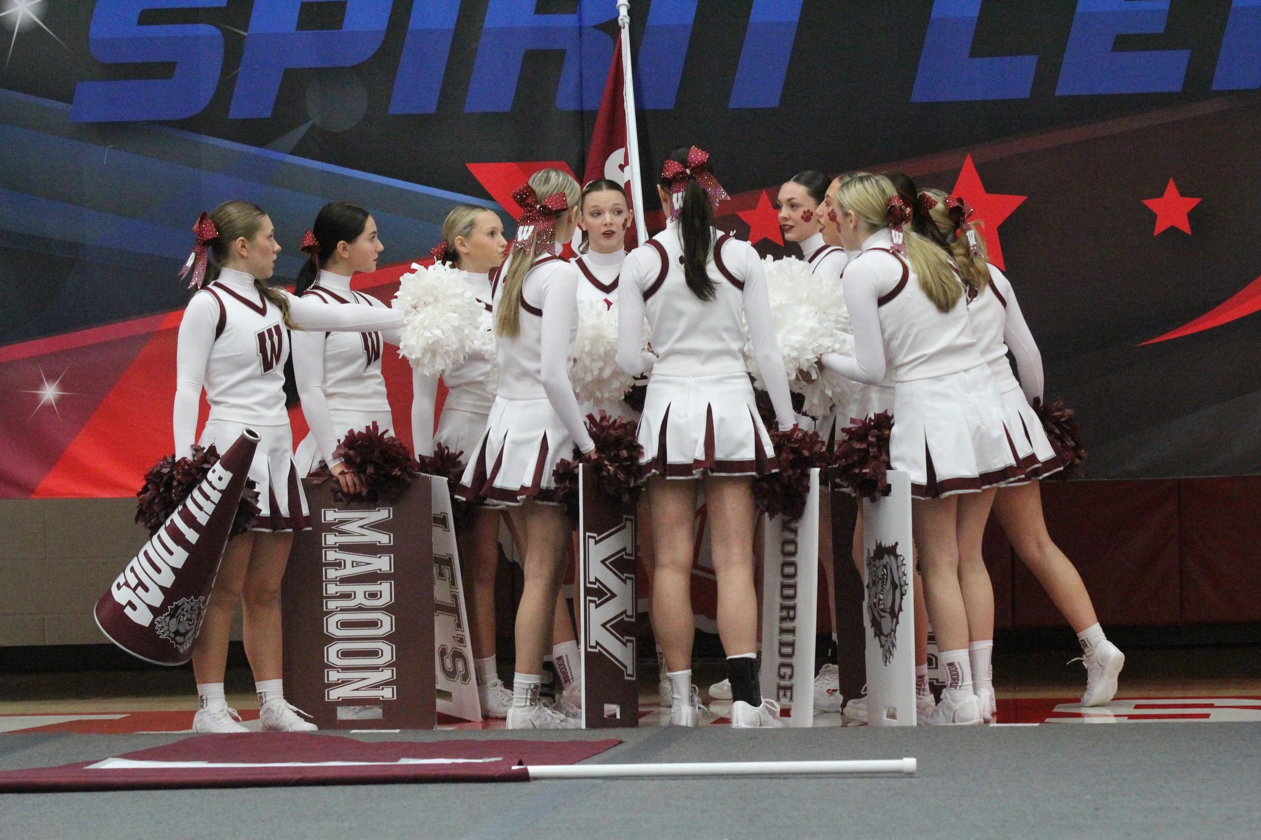 Cheerleaders in white uniforms gather in a huddle holding maroon and white signs and pom-poms.