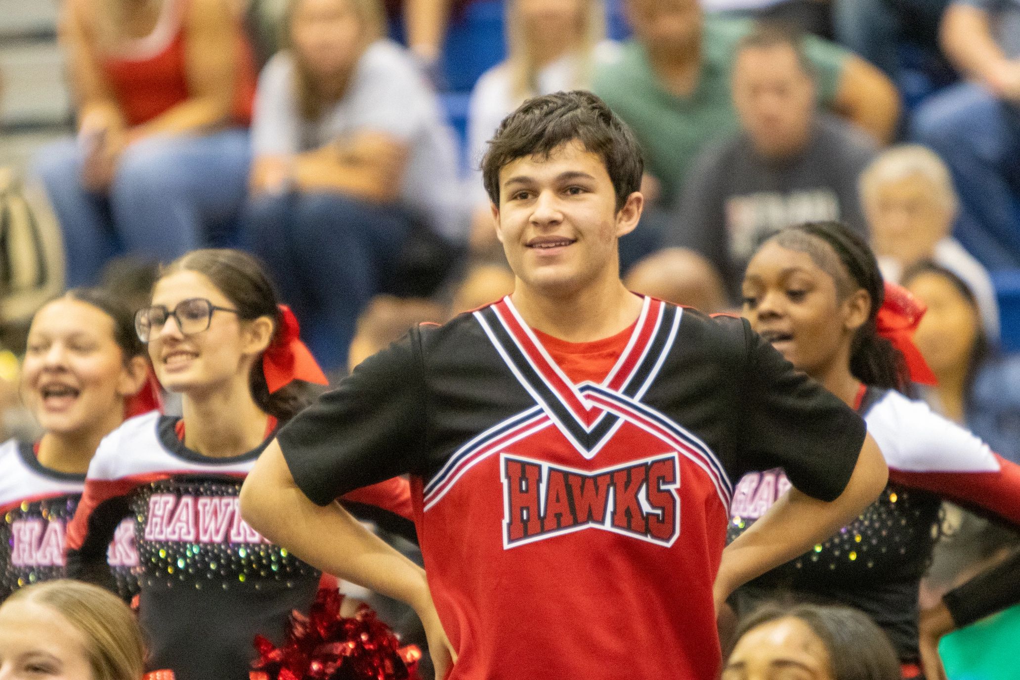 A male cheerleader in a red and black Hawks uniform smiling with teammates behind him.