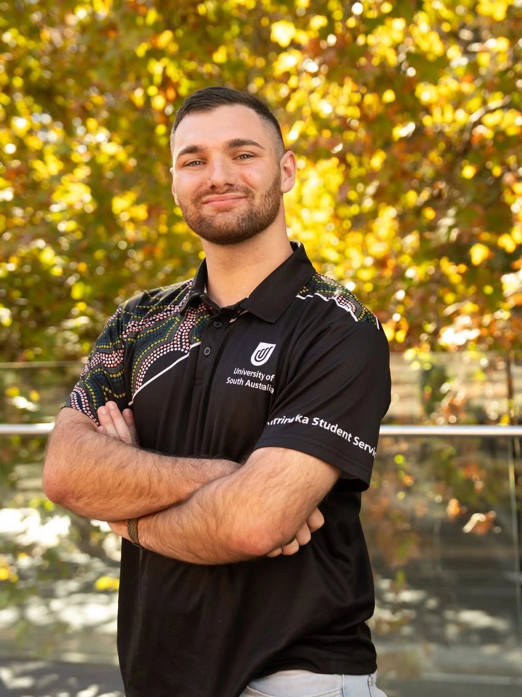Smiling young man in a University of South Australia shirt with crossed arms outdoors.