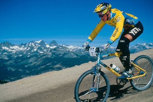 Cyclist in yellow racing on a mountain trail with snowy peaks in the background.