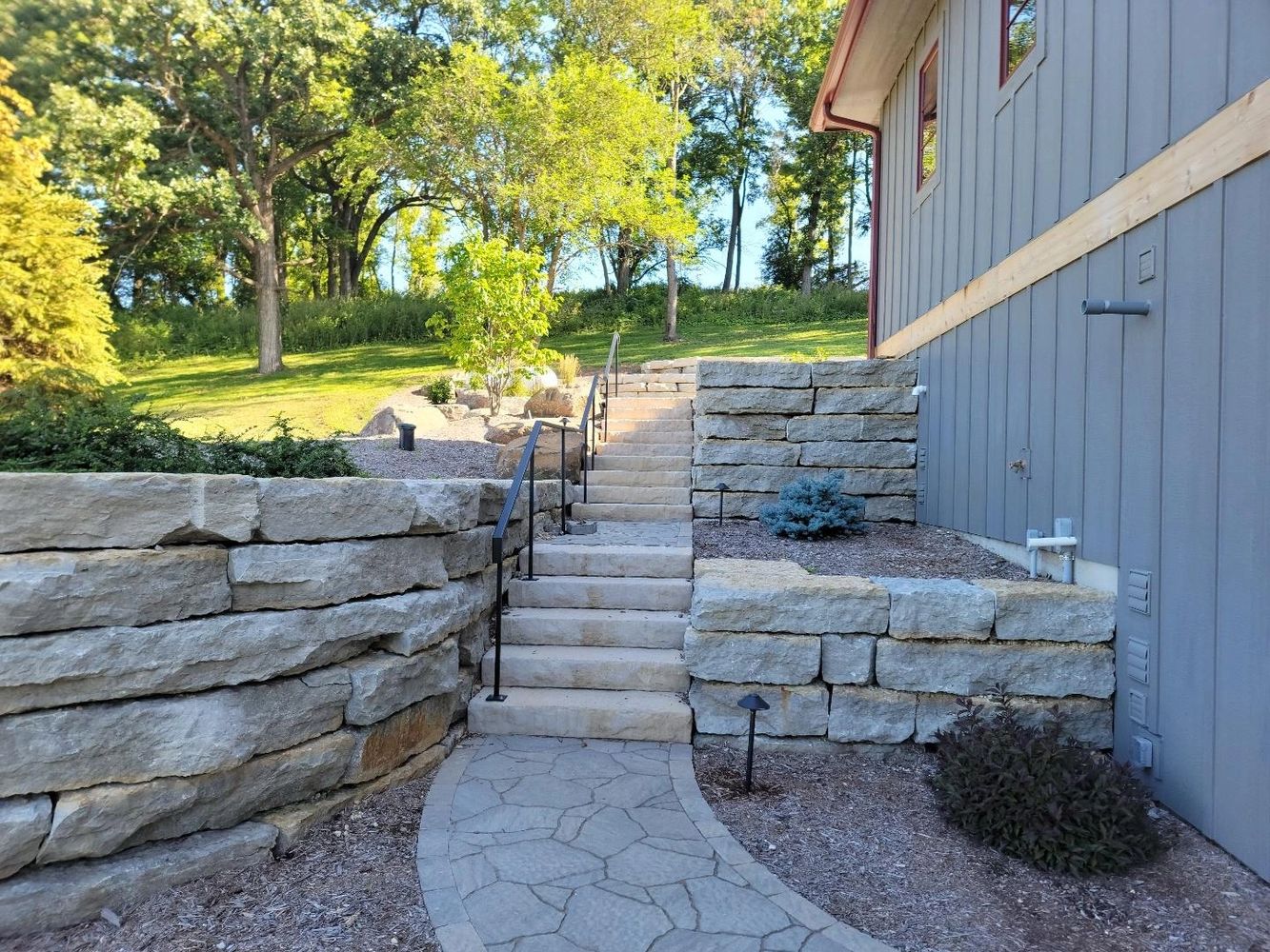 Stone steps with metal railing leading up a landscaped garden beside a gray building.
