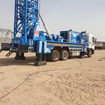 Blue drilling rig mounted on a truck in a sandy area.