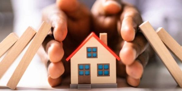 Hands protecting a small wooden house from falling blocks.