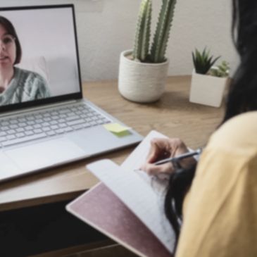 Virtual meeting, one person on the laptop screen and one person at a desk taking notes