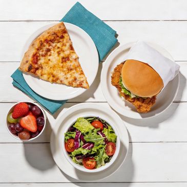 Slice of cheese pizza, chicken sandwich, salad, and fruit cup on white plates.