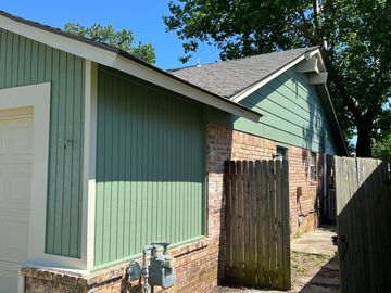 Side view of a green house with brick base and wooden fence under a clear blue sky.