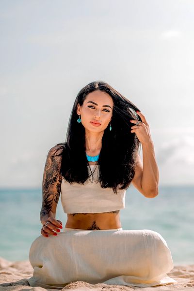 Woman with tattoos sitting on the beach, wearing white and turquoise jewelry.