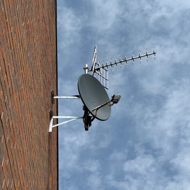 Satellite dish and antenna mounted on a brick wall against a cloudy sky.