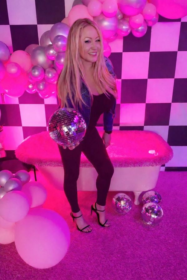 Woman in black outfit holding a disco ball amid pink and silver balloons.