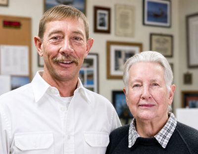 Two older adults posing indoors with framed pictures in the background.