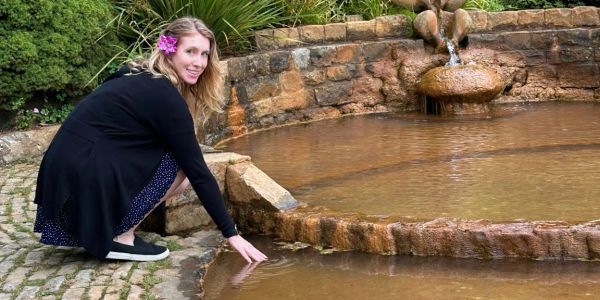 Woman with flower in hair touching water near stone fountain.