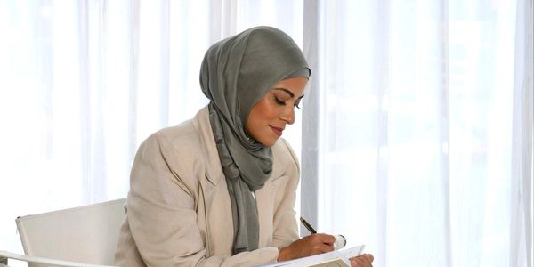 Woman in beige suit and hijab writing in a notebook, seated on a modern chair.