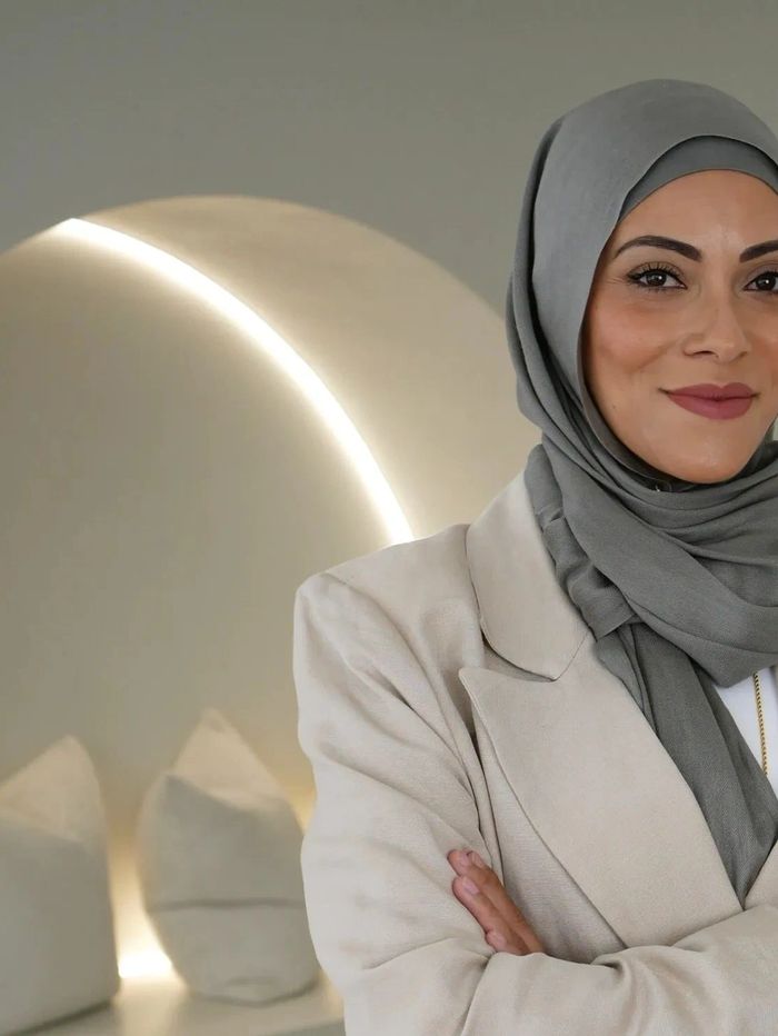 Confident woman in a beige suit and gray hijab standing with arms crossed.