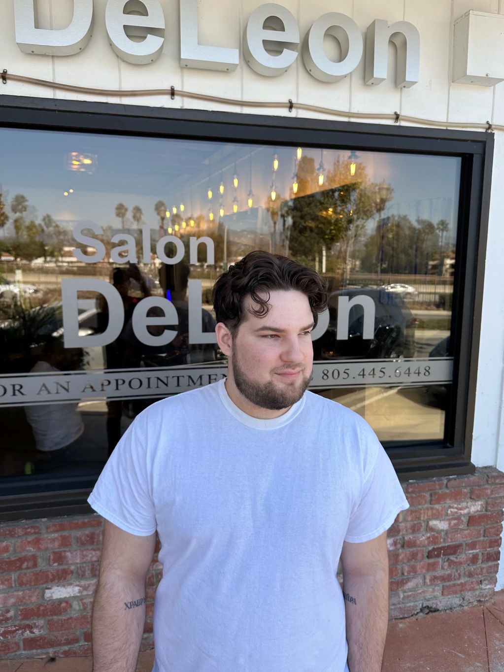 A man in a white t-shirt stands outside Salon DeLeon with a slight smile.
