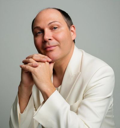 A man in a white suit with hands clasped, smiling gently.