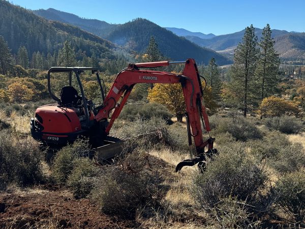 A red Kubota excavator in a mountainous forest clearing under a clear blue sky.