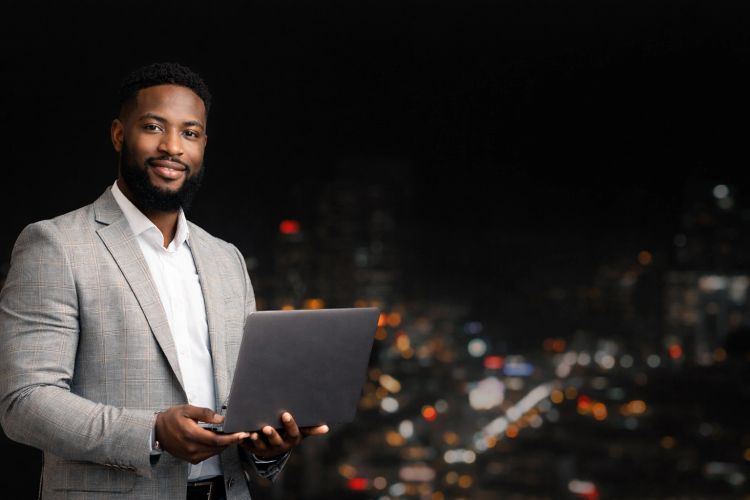 Confident man in a suit holding a laptop with city lights in the background.