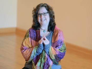 Smiling woman with glasses wearing a colorful shawl, seated indoors.