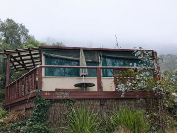 Wooden cabin with a patio umbrella surrounded by greenery and misty hills.
