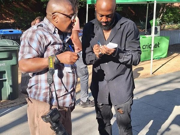 Two men engaged in a conversation outdoors, one holding a camera, the other writing on a notepad.