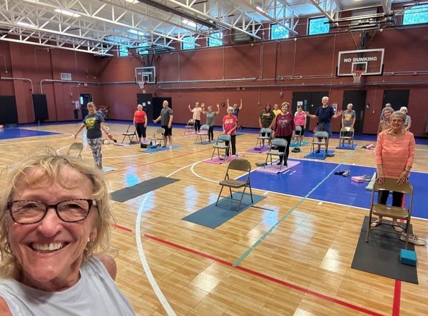 A group of seniors in a gym doing chair yoga or exercise with smiles.