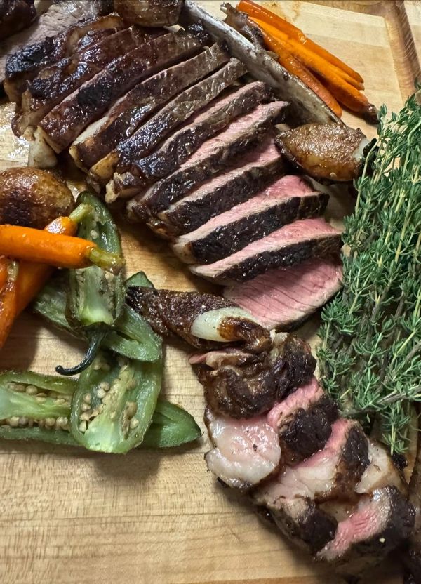 close up of sliced ribeye steak on a wooden cutting board with carrots, grilled jalepeno peppers