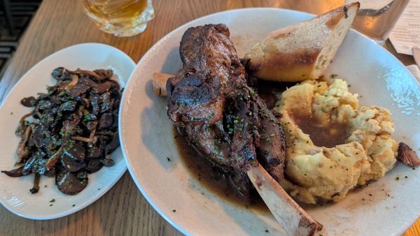 braised lamb shank shown with mashed potatoes and baguette with a side plate of sauteed mushrooms