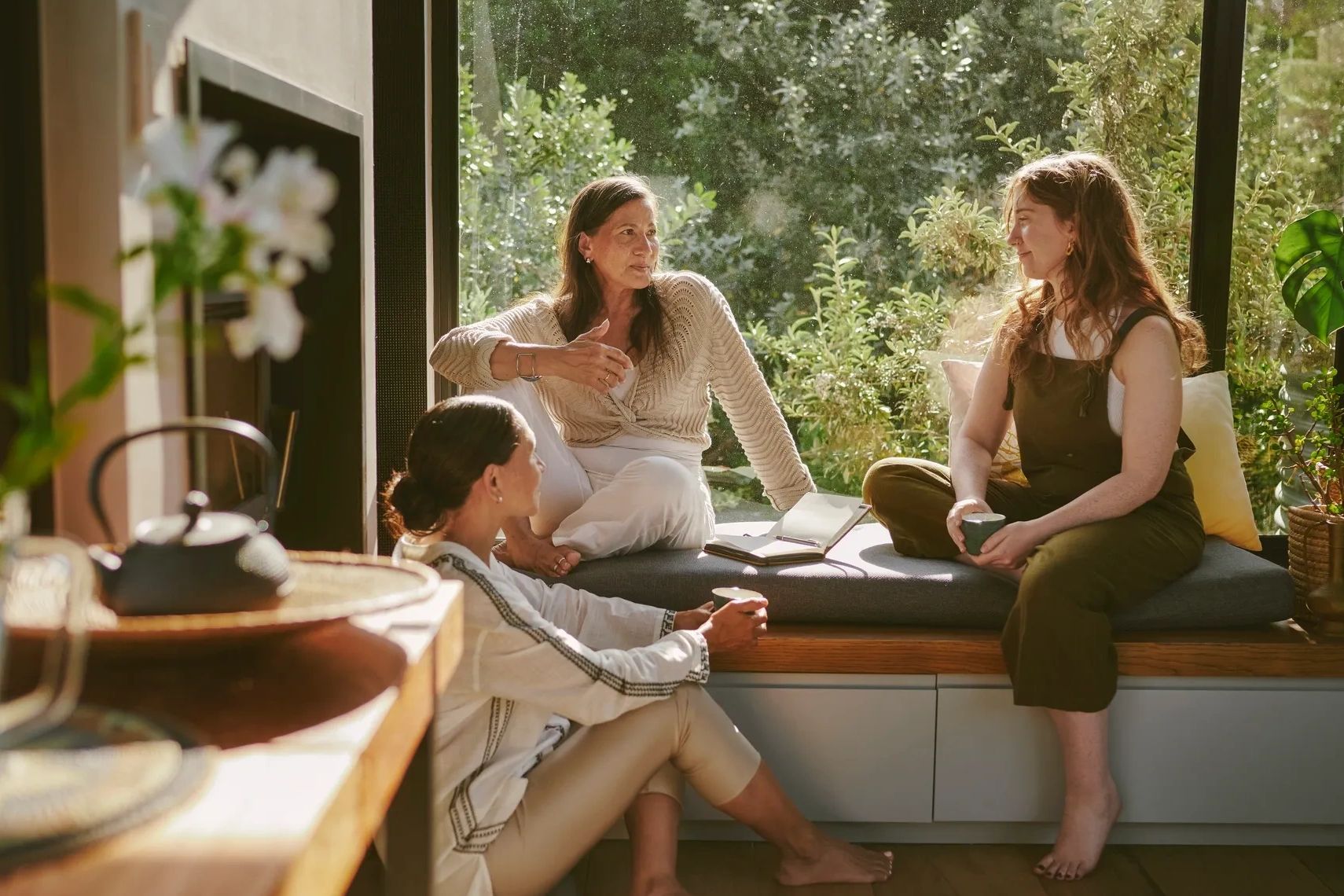 Three women having a relaxed conversation by a large window with greenery outside.