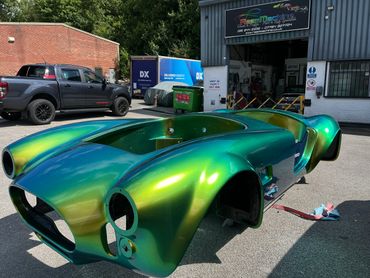 AC Cobra replica car, painted in chameleon custom paint, parked outside the business doors.