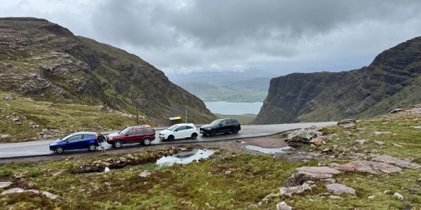 Four cars parked on a mountainous road with a lake in the distance under cloudy skies.