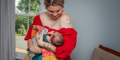 A woman in a red dress tenderly cradling a newborn baby wrapped in a colorful blanket.