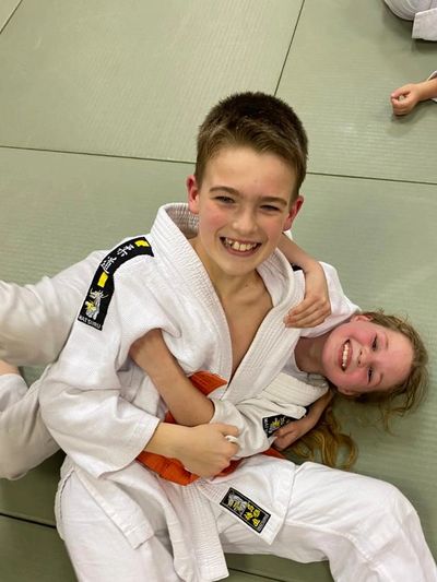 Two kids smiling in judo uniforms on a mat.