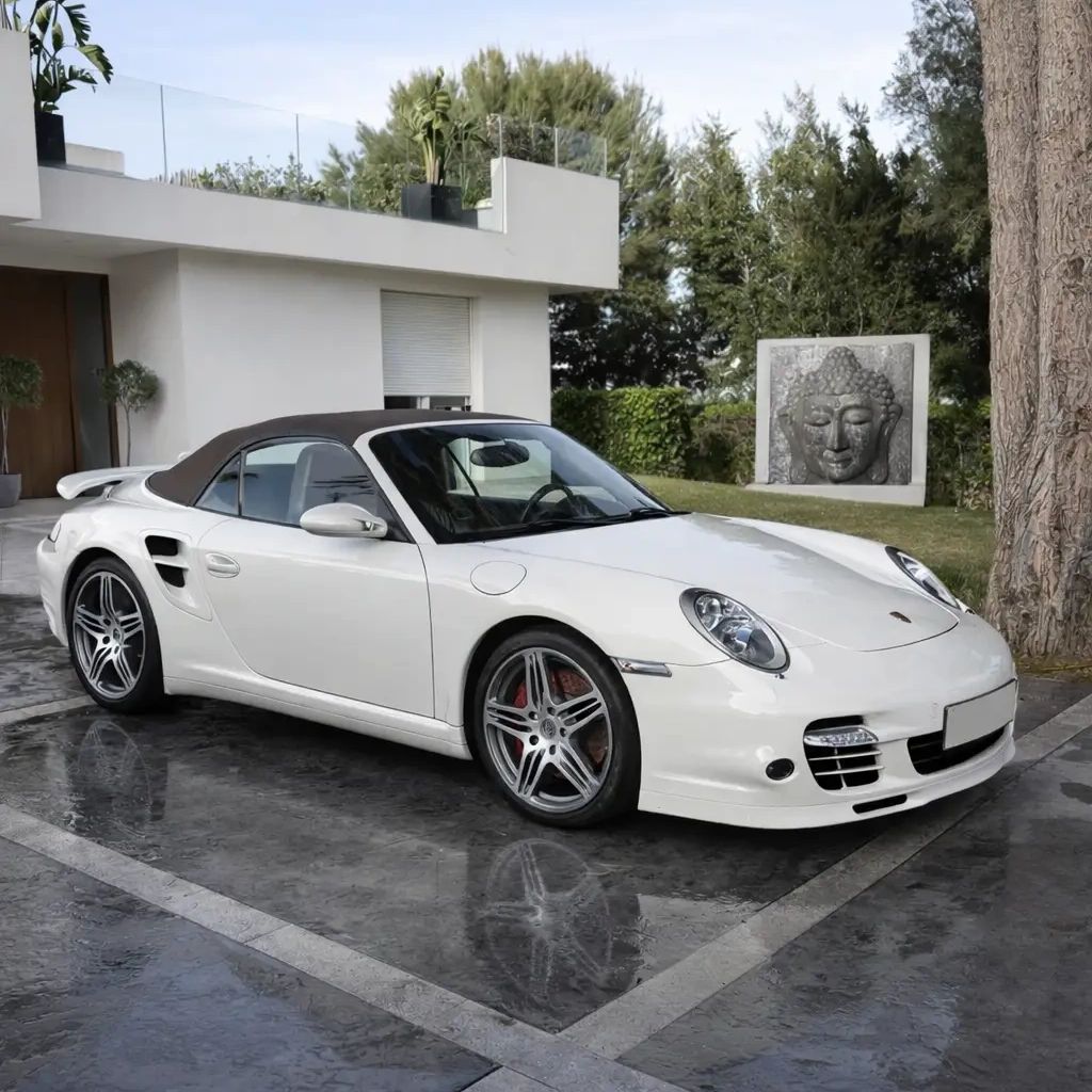 White Porsche convertible parked in a modern driveway with greenery and a Buddha sculpture.