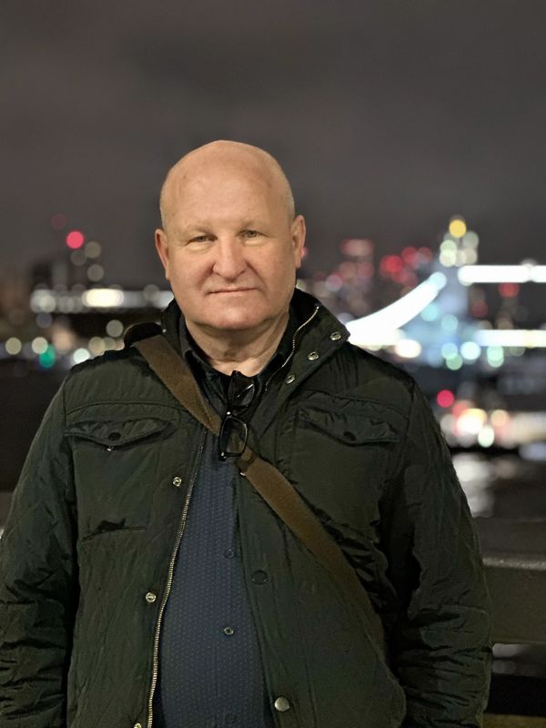 Man in dark jacket posing at night with city lights blurred in the background.