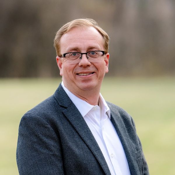 Smiling man in glasses and a gray blazer outdoors.
