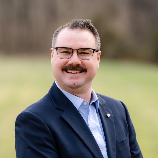 Smiling man with glasses and mustache in a blue suit outdoors.