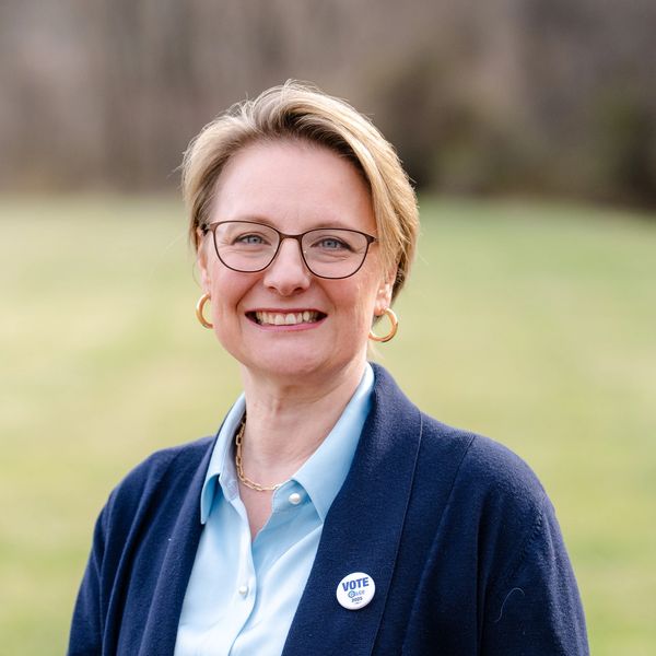 Smiling woman wearing glasses and a 'Vote' pin outdoors.