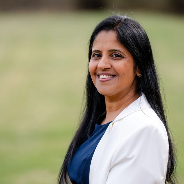 A smiling woman with long black hair, wearing a white blazer and blue top, outdoors.