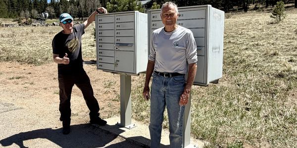 Two men standing next to outdoor community mailboxes on a sunny day.