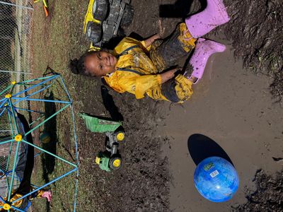 Child in yellow raincoat and purple boots playing happily in muddy puddle.