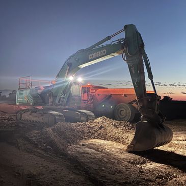 Excavator at dusk on a construction site with lights on.