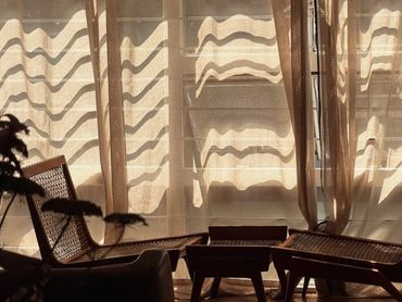 Luz del sol filtrándose a través de cortinas frente a sillas de mimbre.