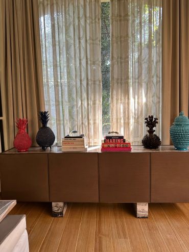 Colorful pineapple sculptures and books decorate a modern brown cabinet in front of sheer curtains.