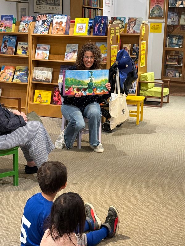 A woman reads a colorful book to children in a cozy library setting.