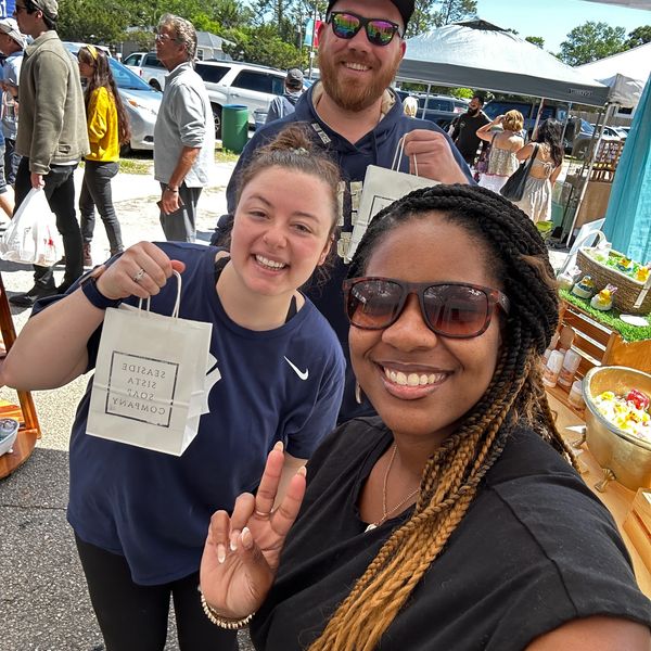 Three friends smiling at an outdoor market, holding shopping bags.